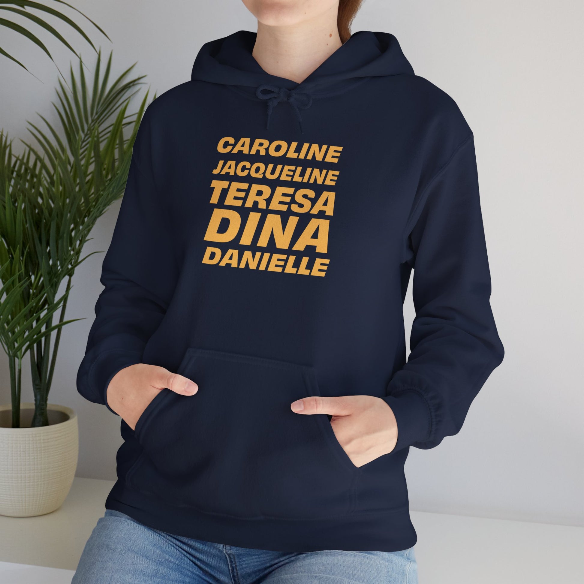 Female model indoors wearing navy OG NJ Housewives Hooded Sweatshirt with hands in pocket, yellow names on chest.