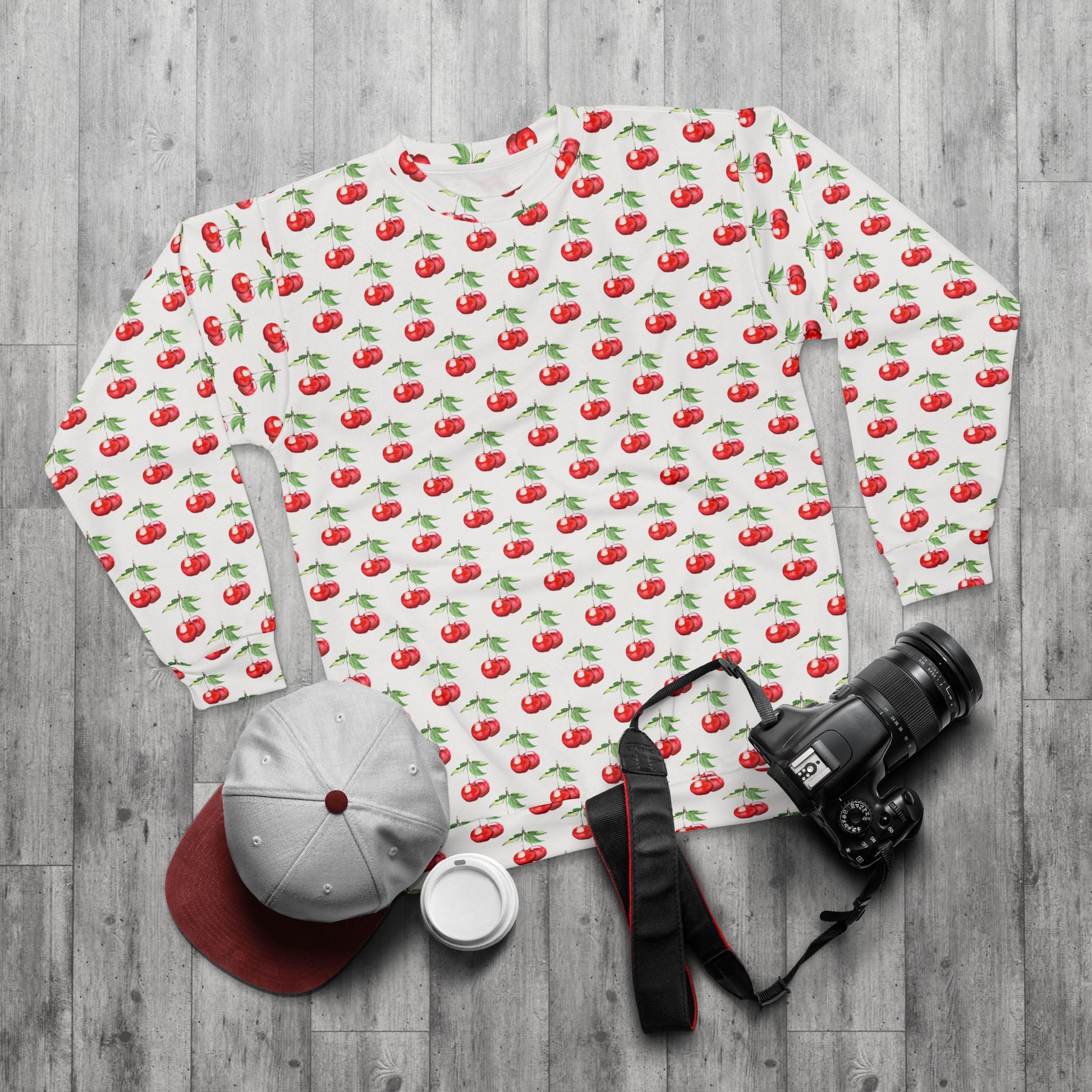 Sweet Cherry Sweatshirt laid out on a wood surface alongside a camera, hat, and mug, emphasizing casual lifestyle appeal.
