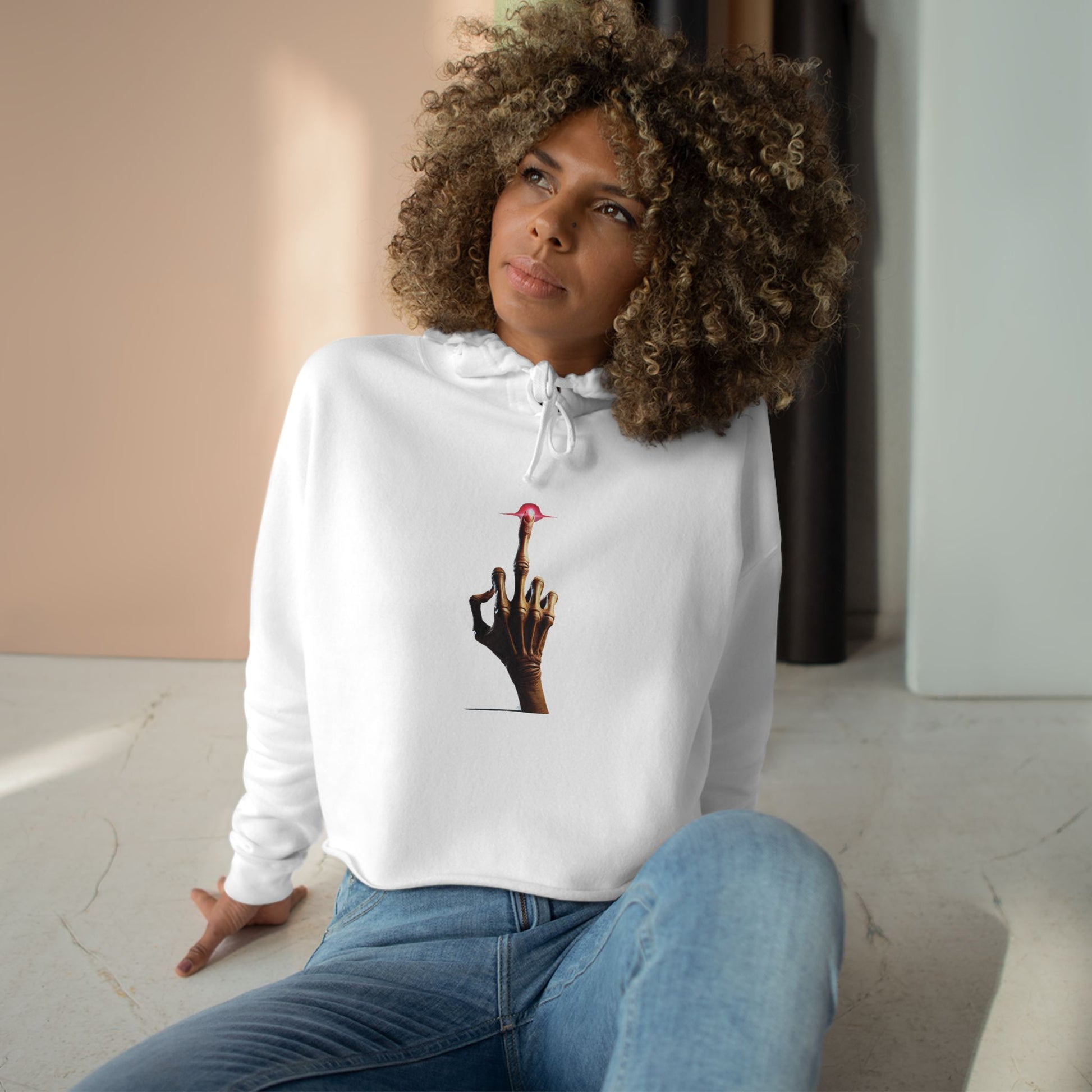 A woman with curly hair sits on the floor in natural light, wearing a white crop hoodie with a minimalist E.T.-inspired graphic of a hand with an extended glowing finger touching a small red spaceship.