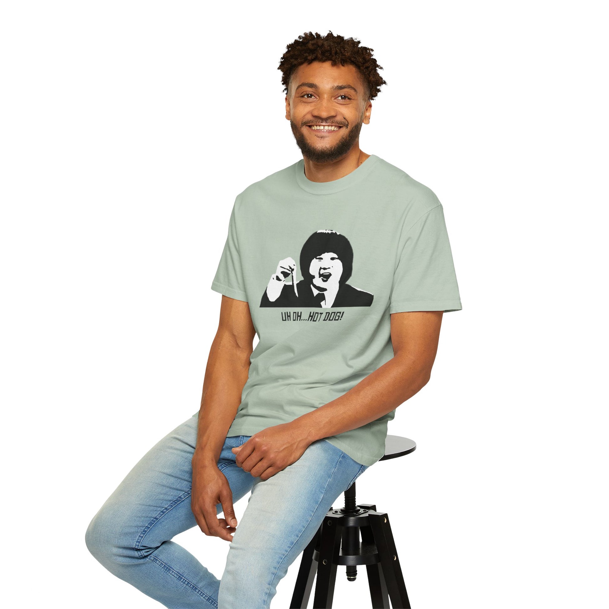 Man sitting on stool wearing light green Uh Oh Hot Dog T-shirt, smiling.