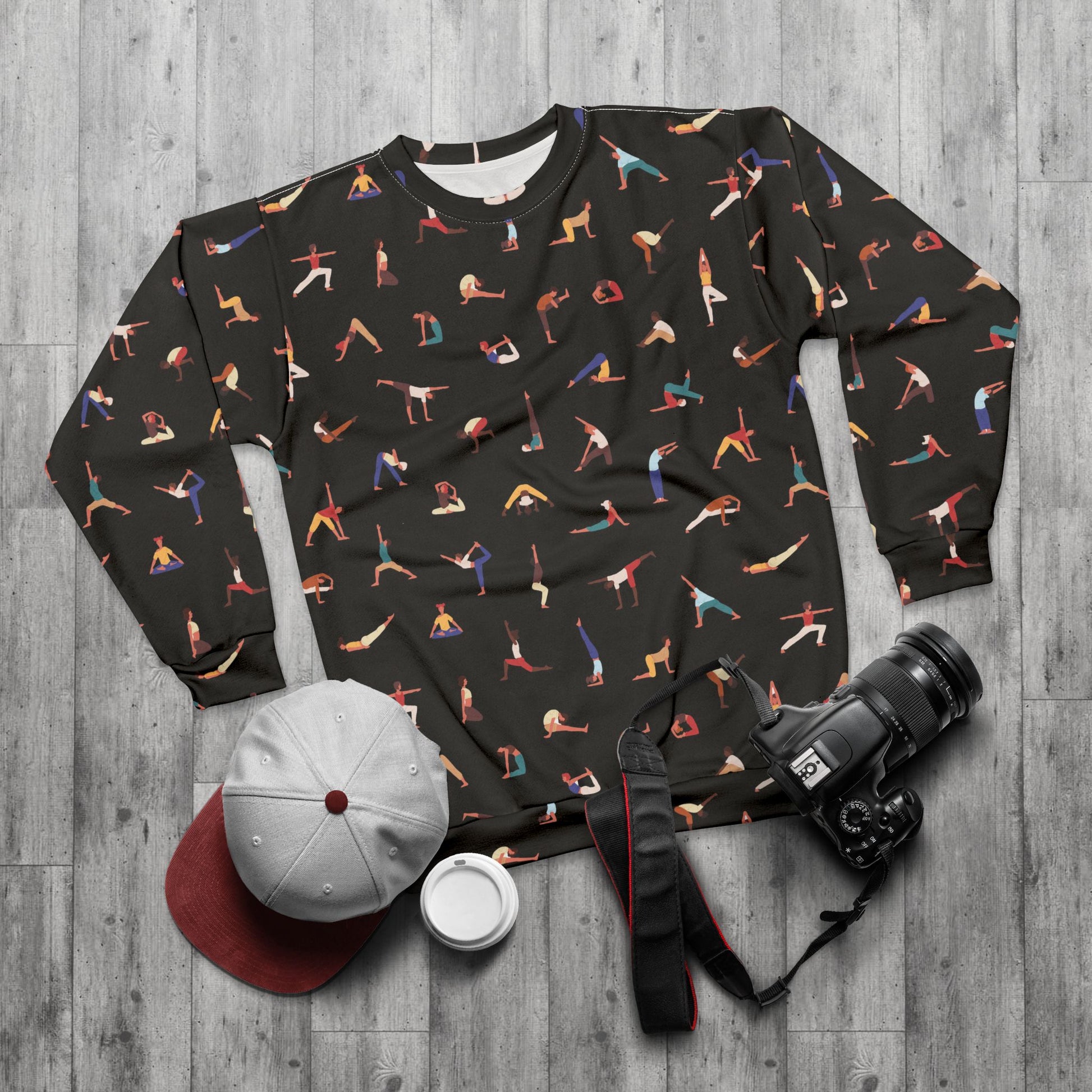 Black sweatshirt with colorful yoga pose pattern on a wooden surface with camera, cap, and cup.