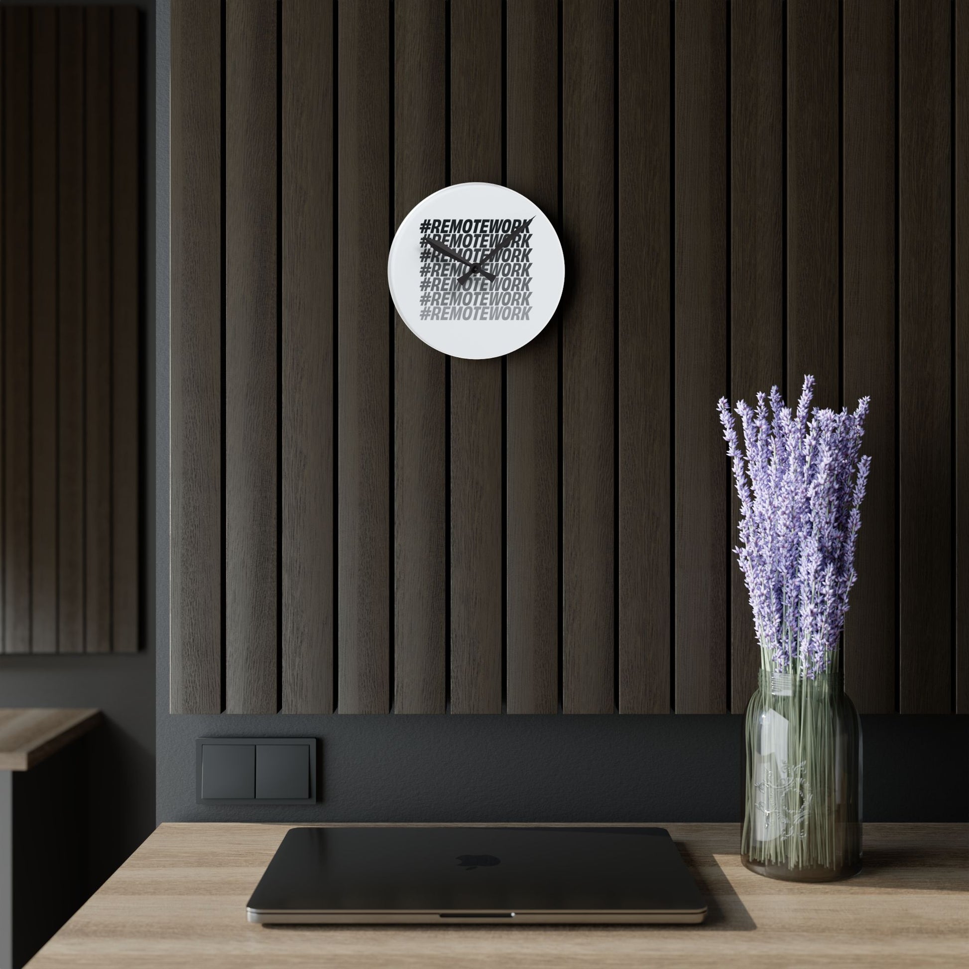 Wall clock with text design on a dark wall with a vase of lavender on a table.
