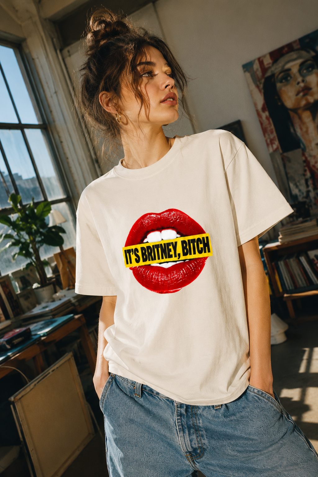 A woman wearing a cream-colored graphic t-shirt with a red lips design and the text 'IT'S BRITNEY, BITCH' in yellow, paired with blue jeans in a sunlit room.