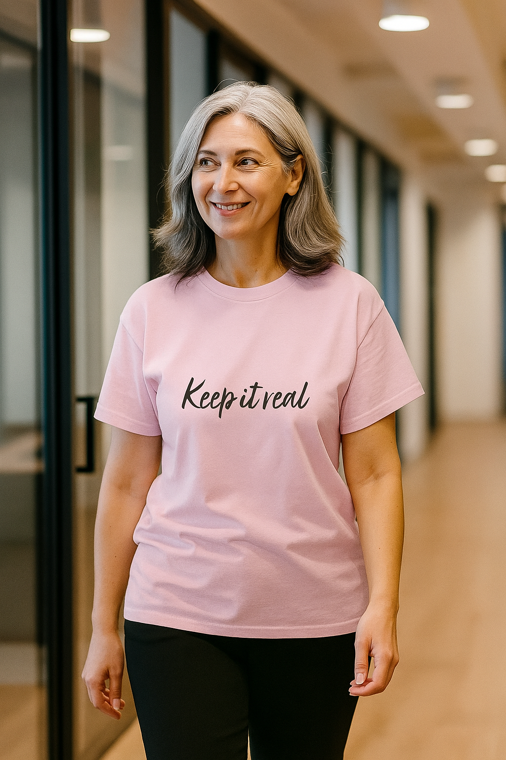 Smiling woman with natural grey hair walking confidently in a hallway while wearing a pink Keep It Real t shirt.