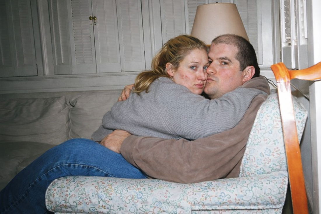 Conceptual “what-if” portrayal inspired by JFK Jr. and Carolyn Bessette, showing a heavier-bodied couple embracing on a couch in an ordinary living room to challenge beauty and status expectations