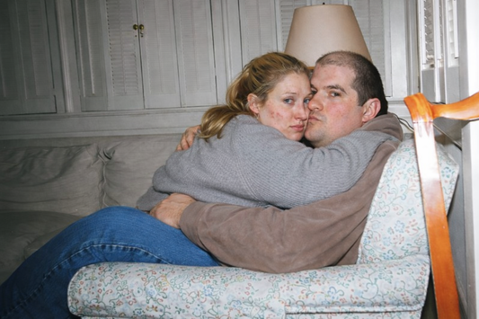 Conceptual “what-if” portrayal inspired by JFK Jr. and Carolyn Bessette, showing a heavier-bodied couple embracing on a couch in an ordinary living room to challenge beauty and status expectations