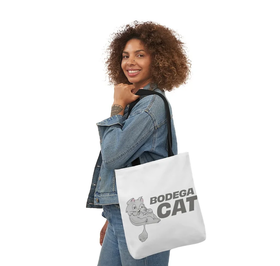 Smiling woman with curly hair wearing a denim jacket and holding a tote bag that reads "Bodega Cat" featuring a cartoon cat design.