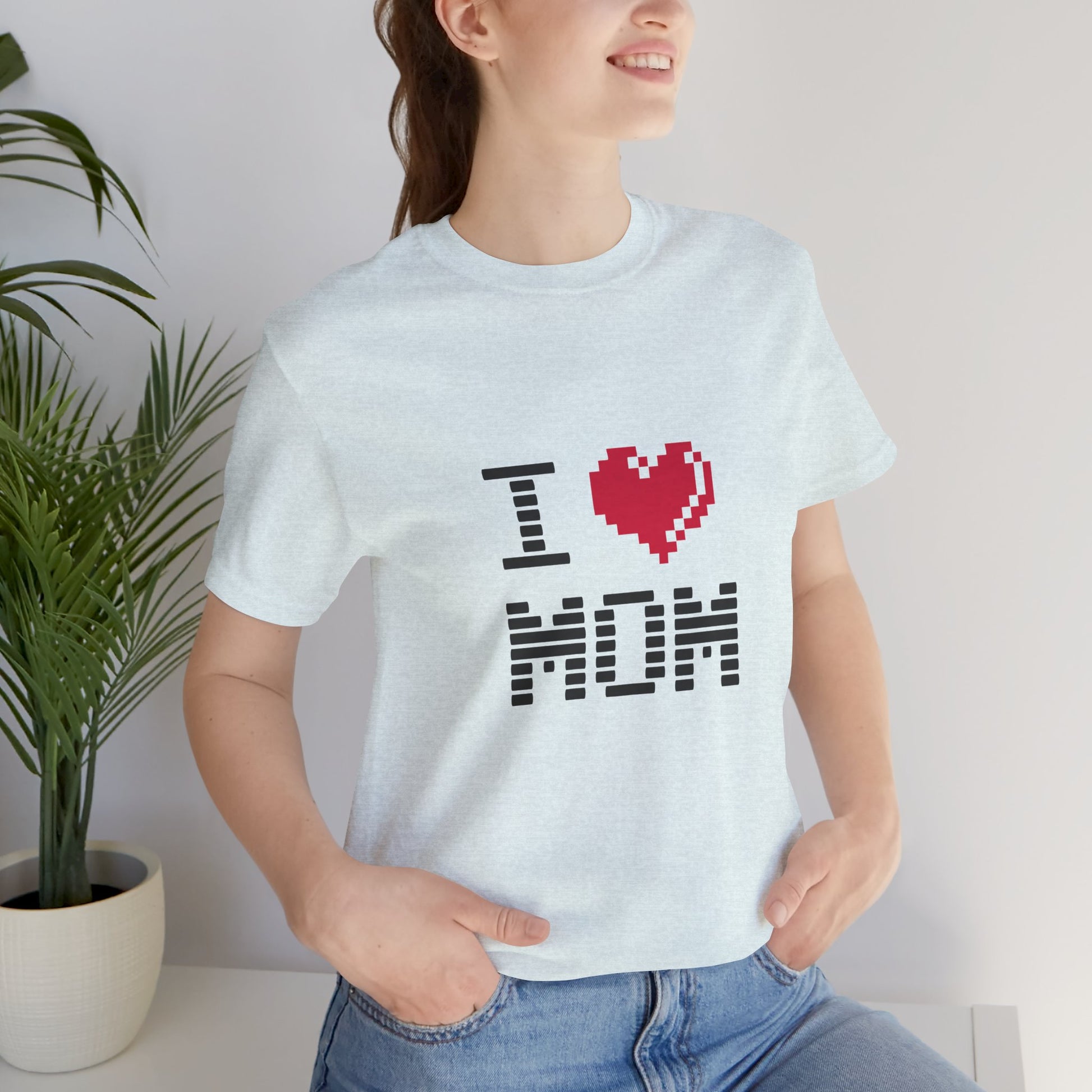 Person wearing a white t-shirt with 'I ❤️ MOM' design, standing indoors next to a plant.
