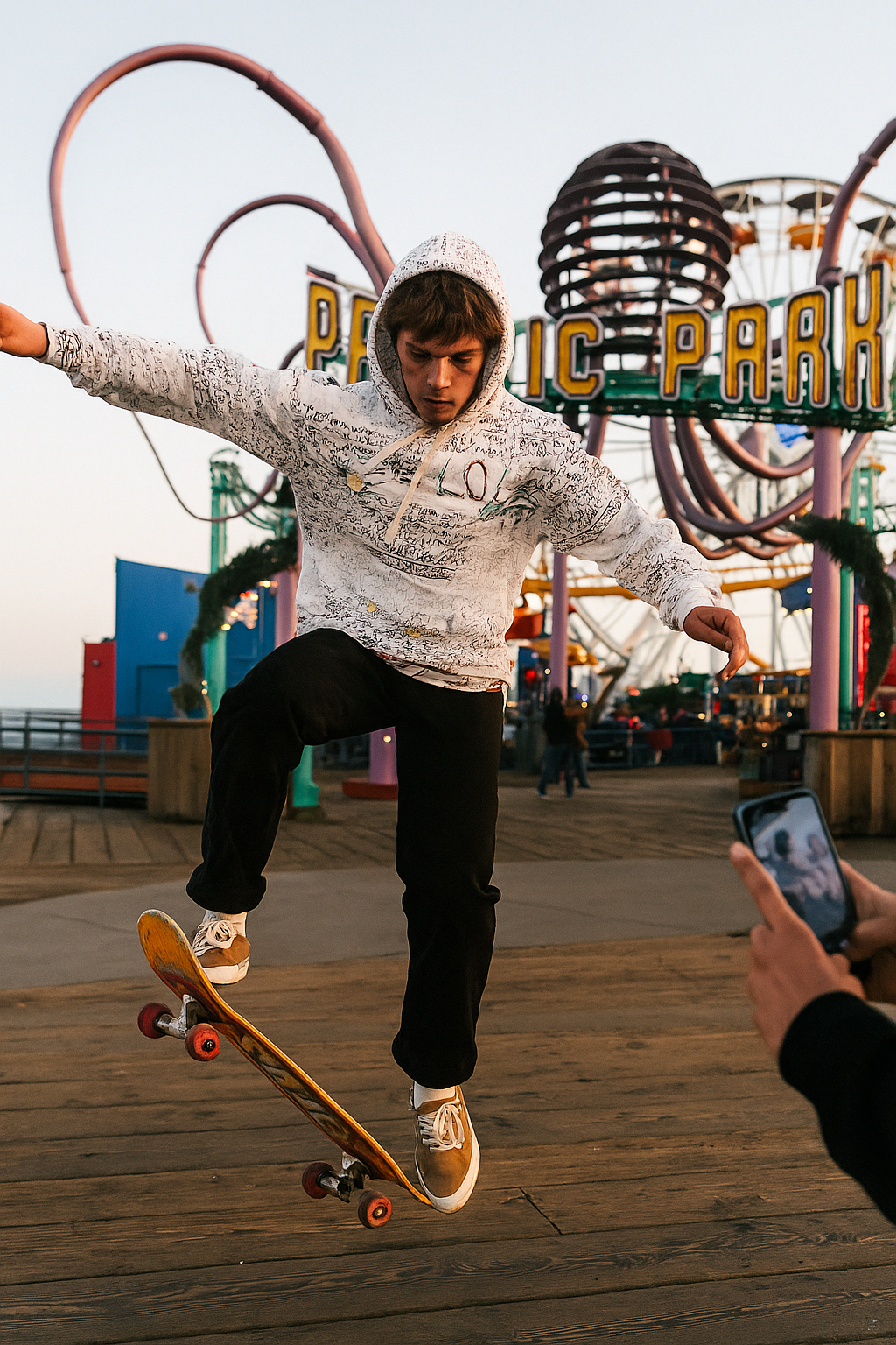 Young skateboarder performing a trick on the Santa Monica Pier while wearing the True Love Hoodie by Tantrum Media Store, with Pacific Park rides glowing in the background.
