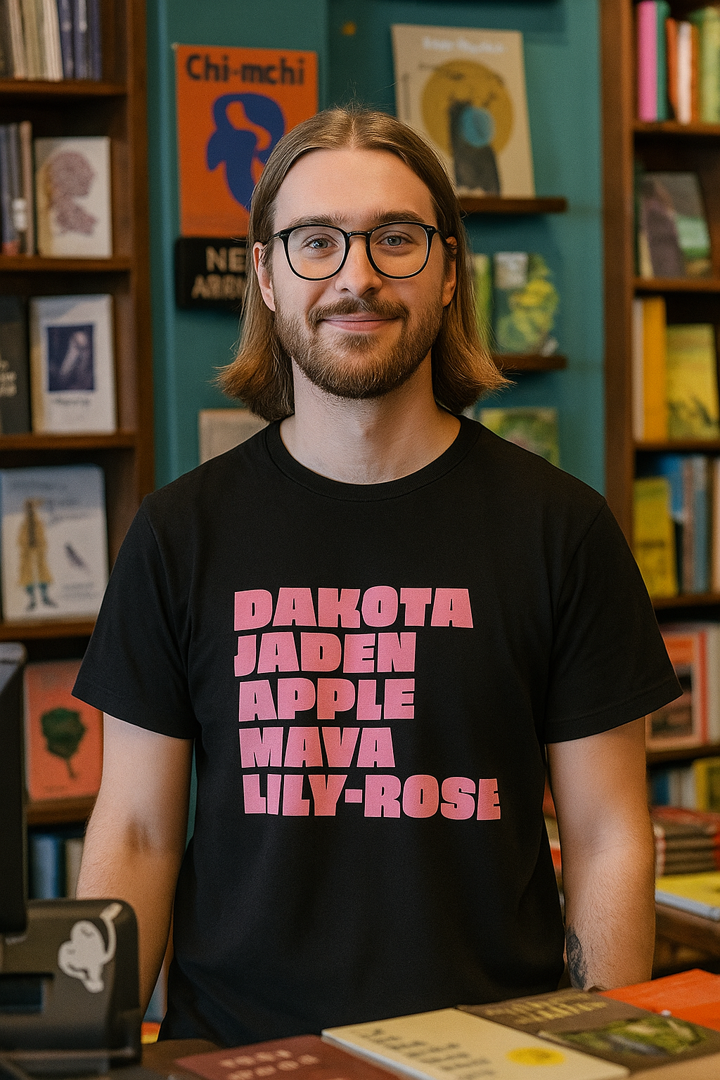 Person standing inside a bookstore wearing a black T-shirt with bold pink text listing names, smiling, with bookshelves and colorful covers in the background.