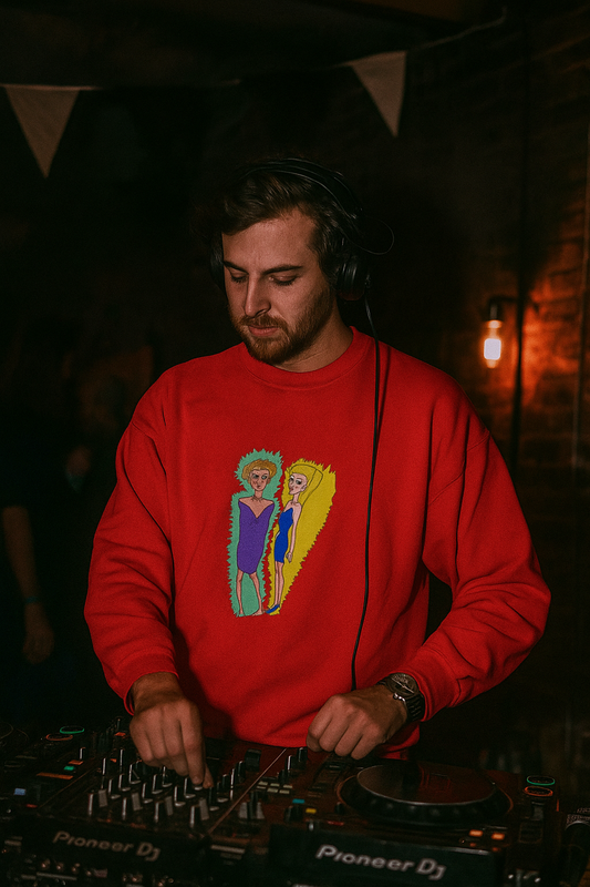 Man DJing in a dimly lit venue while wearing a bright red sweatshirt featuring a colorful hand drawn illustration of two abstract characters.