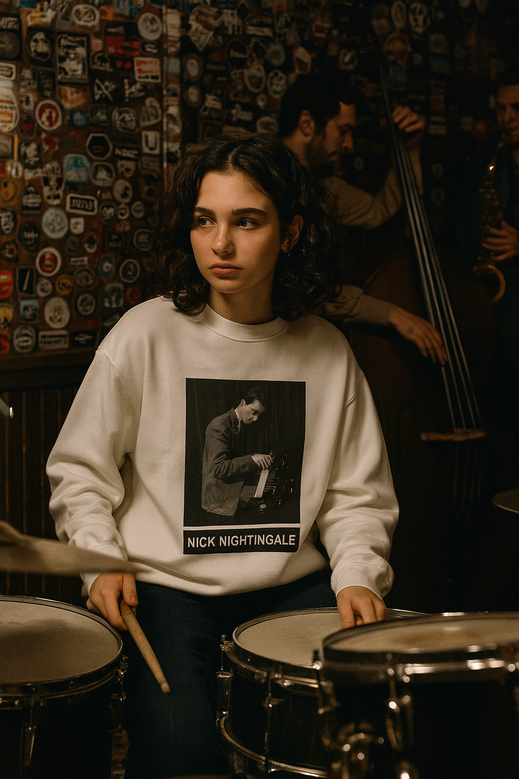 A young person sitting at a drum kit wearing a white Nick Nightingale sweatshirt in a dimly lit music space.