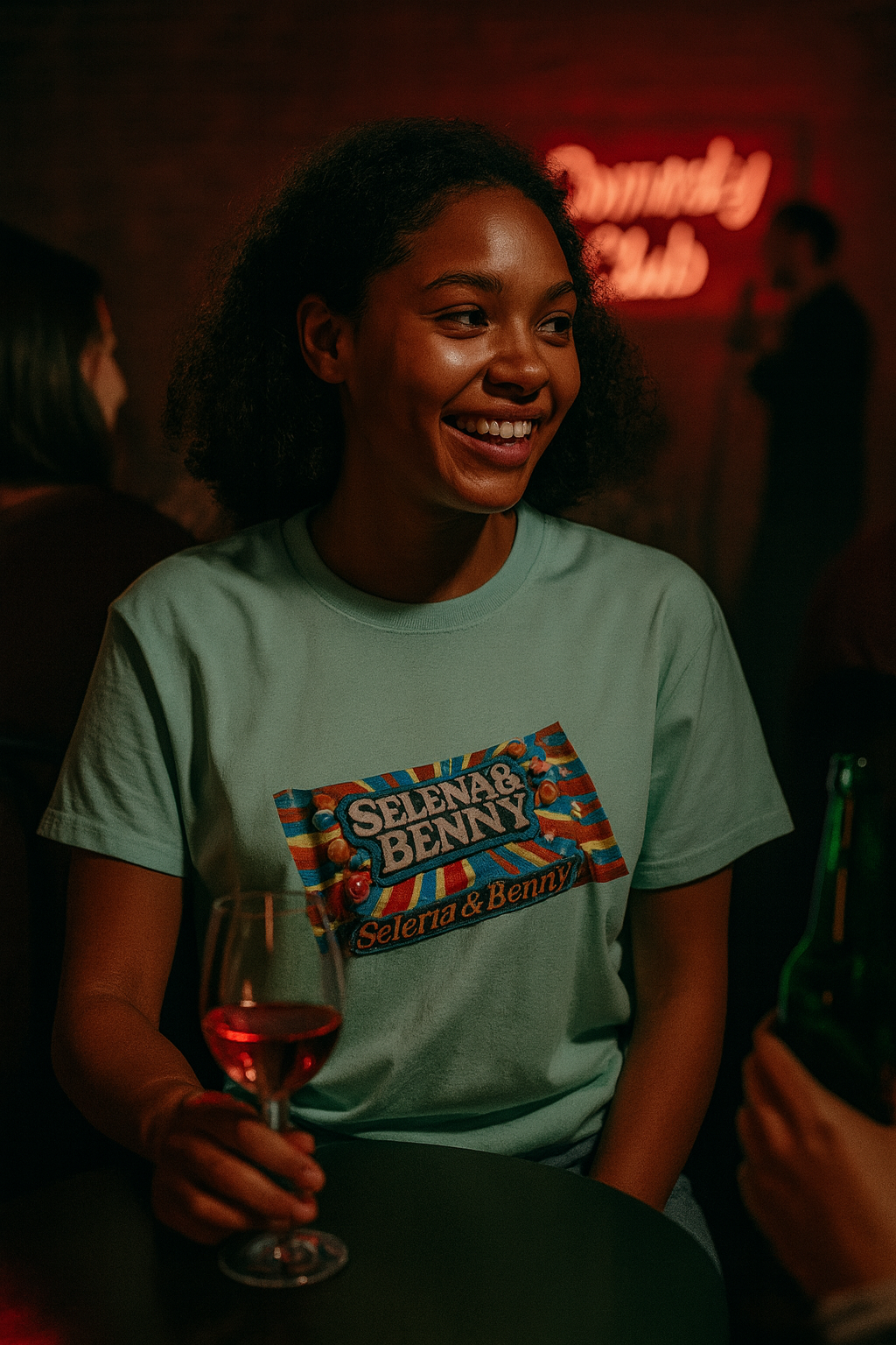 Person wearing a 'Selena & Benny' t-shirt in a dimly lit bar setting.