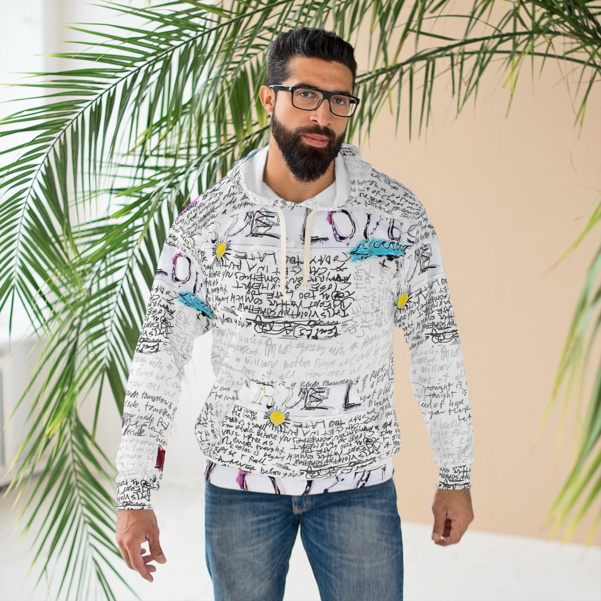 Male model wearing white unisex hoodie with handwritten text and doodle artwork, posing outdoors near palm leaves
