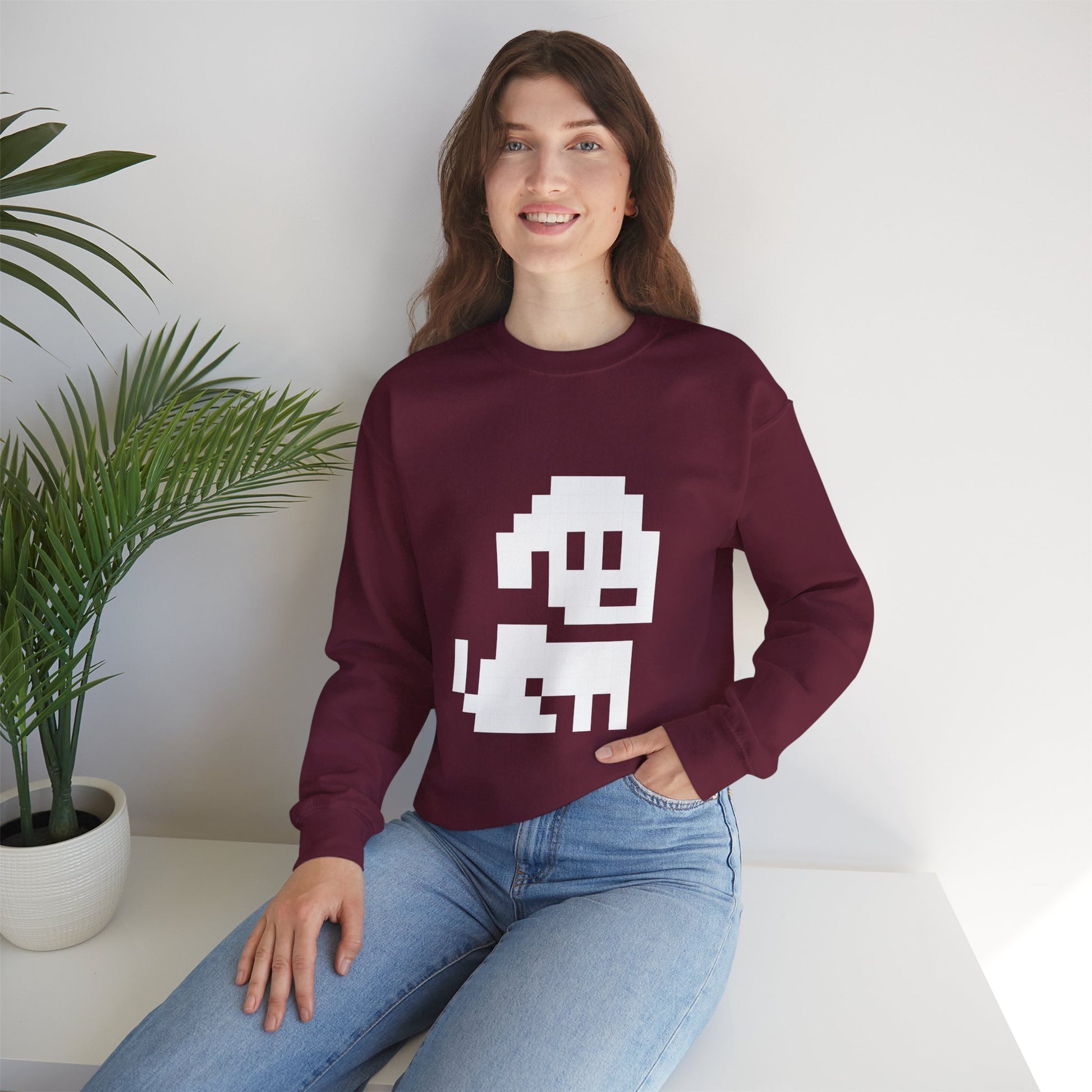 Woman wearing a maroon sweatshirt with a white graphic design, sitting on a white surface with a plant in the background.