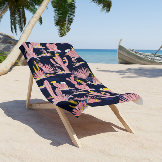 Beach scene with pink cactus patterned towel draped over a wooden lounge chair on sand near palm trees and ocean