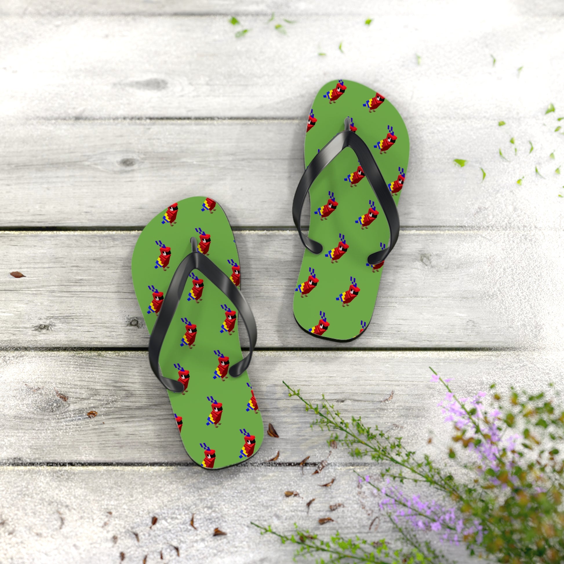 Pair of green flip flops with red arcade parrot pattern and black straps displayed on wooden surface with plants