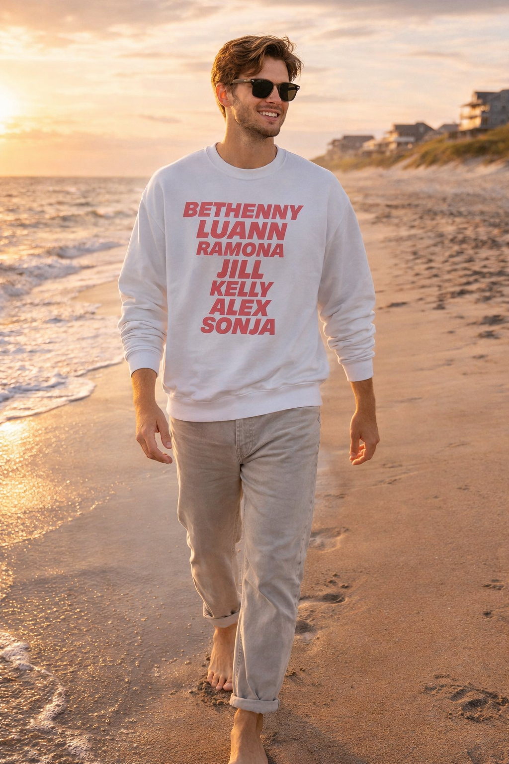 Man walking on a beach wearing white OG NY Housewives men’s sweatshirt with stacked cast name graphic