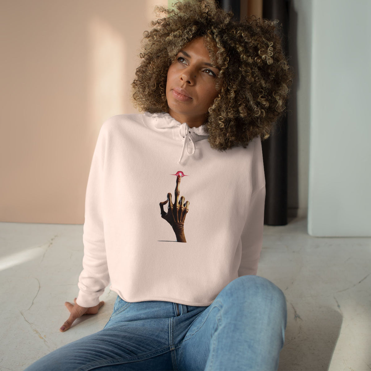 A woman with curly hair sits on the floor in natural light, wearing a light pink crop hoodie with a minimalist E.T.-inspired graphic of a hand with an extended glowing finger touching a small red spaceship.