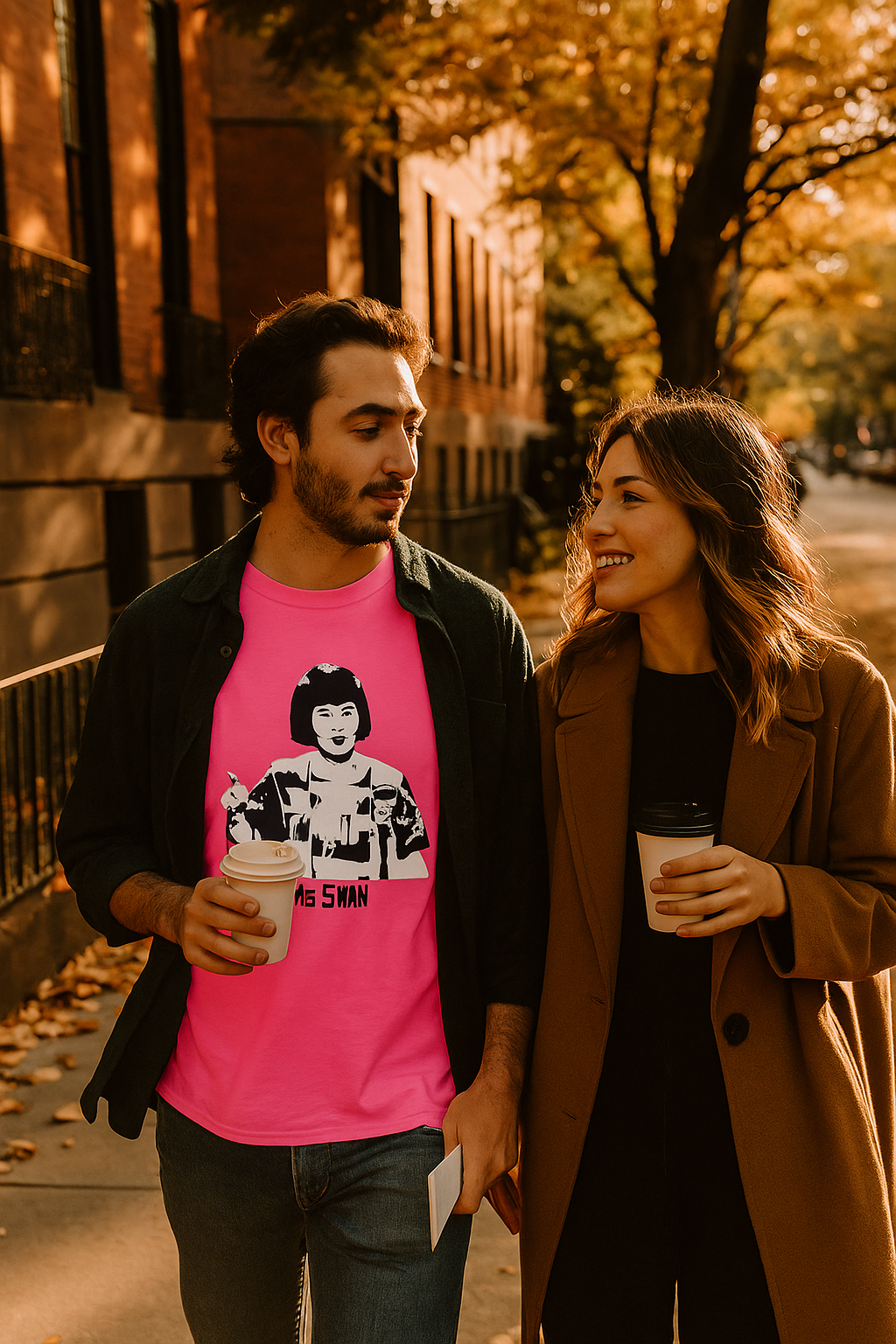 Man and woman walking together on a street, holding coffee cups, with autumn leaves and buildings in the background. He is wearing a hot pink Ms. Swan Unisex T-Shirt