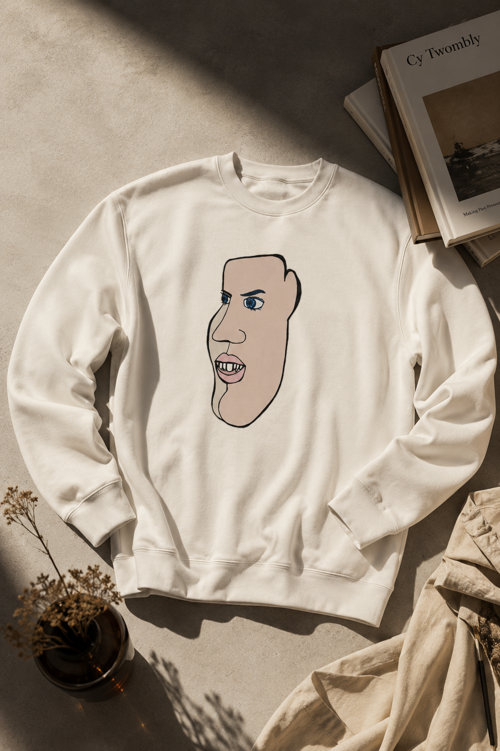 Soft cream oversized crewneck sweatshirt with abstract face illustration styled in a warm editorial flat lay with gradient neutral lighting, artistic books, and minimalist studio props for a cozy contemporary aesthetic.