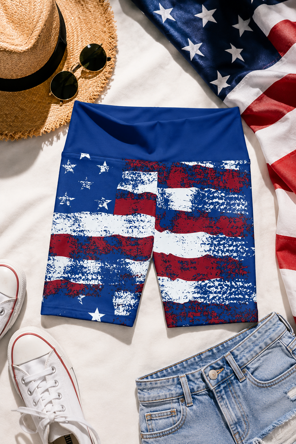 Editorial flat-lay photography of distressed American flag bike shorts styled with a straw hat, sunglasses, white sneakers, denim shorts, and patriotic fabric on a bright summer background.
