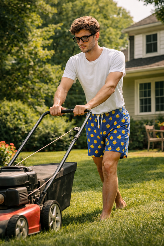 Gen Z man mowing the lawn in a suburban backyard wearing blue duck-print swim shorts and a white t-shirt on a sunny day.