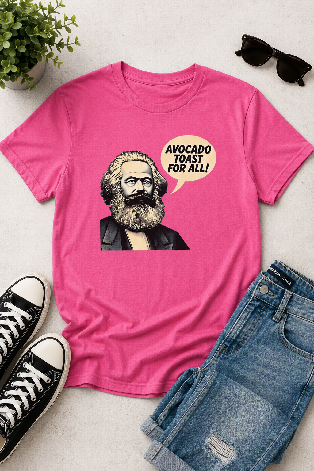 Pink graphic statement t-shirt laid flat with jeans, sneakers, and sunglasses, featuring Karl Marx artwork and the slogan “Avocado Toast for All.”