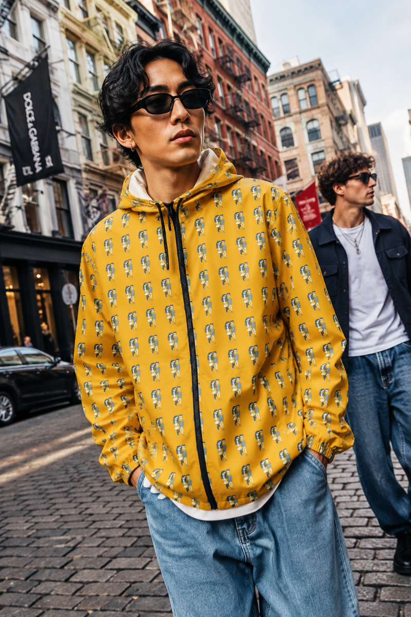 A man wearing a Babes,  bright yellow zip-up hoodie with a repeating black and white graphic pattern, styled with blue jeans and sunglasses, standing on a cobblestone city street.