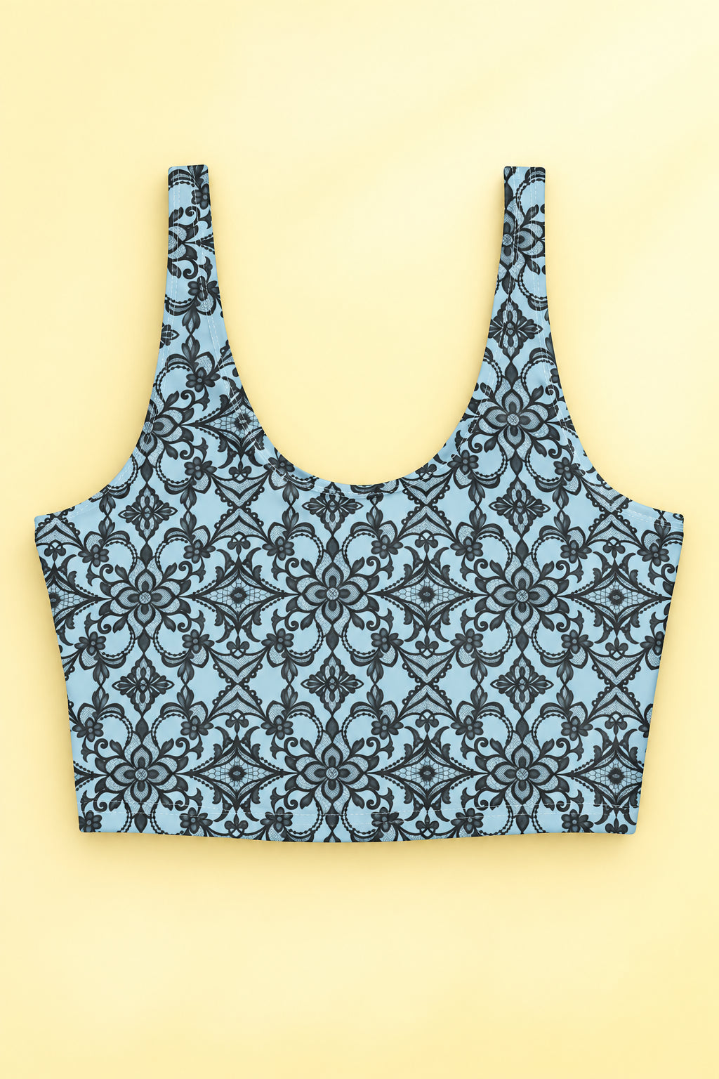 Front flat lay of blue sports bra with intricate black floral lace print, wide shoulder straps and scoop neckline on a pastel yellow background.