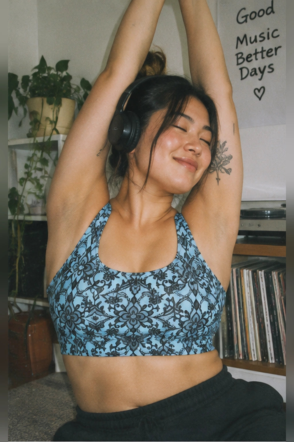 Woman wearing a blue floral lace print sports bra and black sweatpants while stretching with headphones in a cozy room with vinyl records.