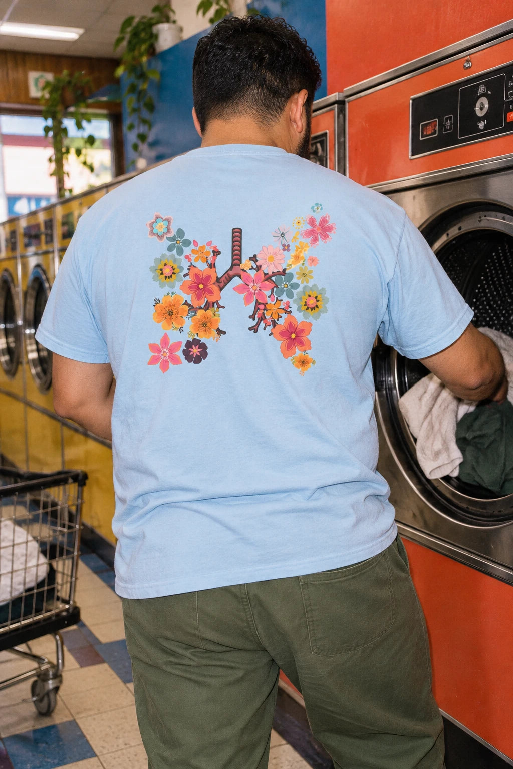 Man loading a washing machine while wearing the light blue Breath T-shirt with floral lungs back print.