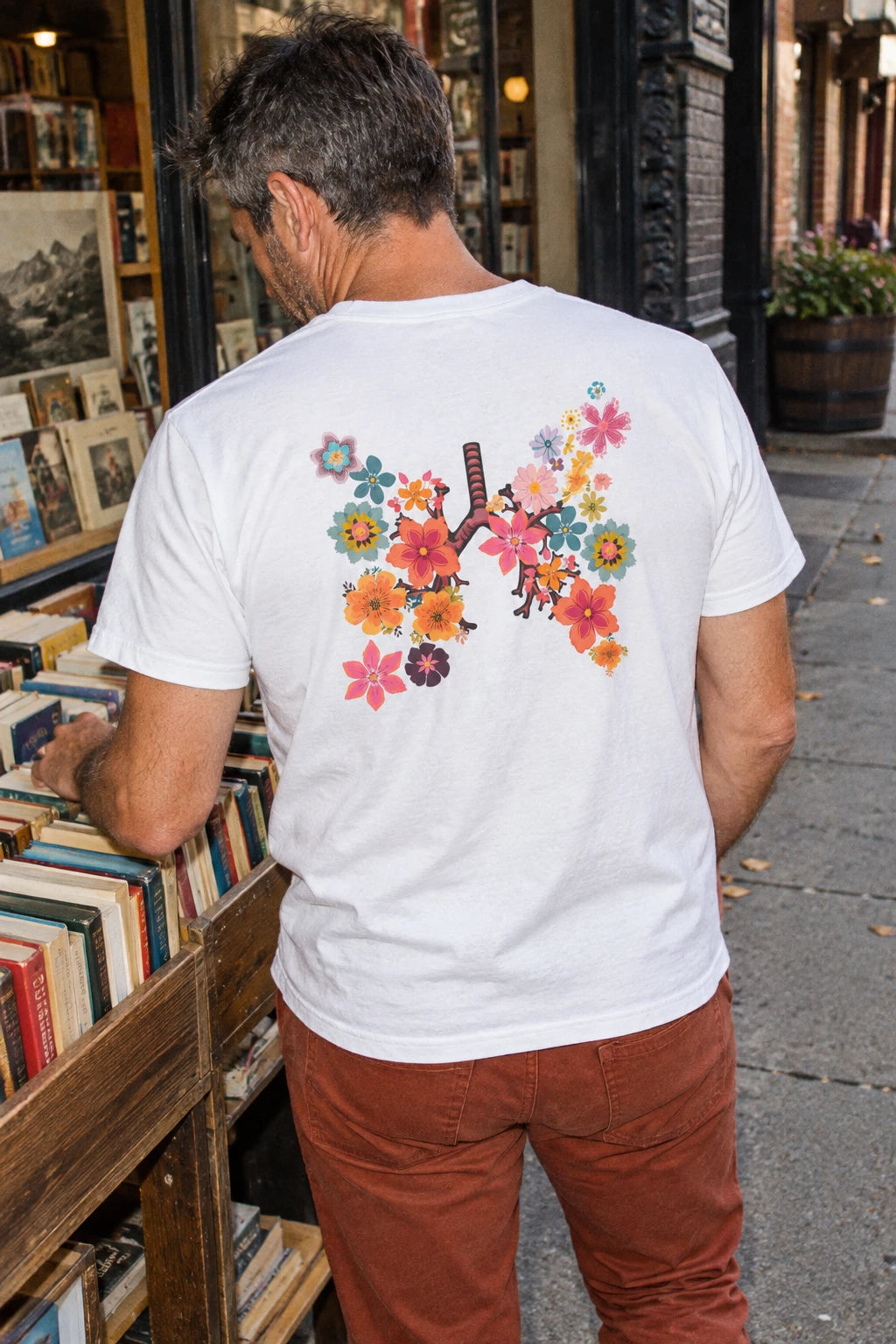 Man wearing the white Breath T-shirt with a colorful floral lungs back print while browsing outdoor books.