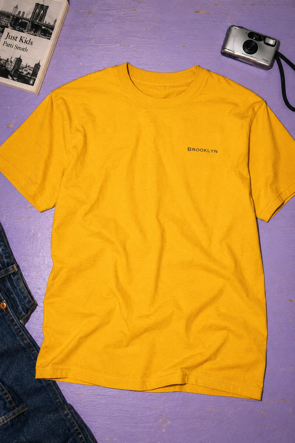 Golden yellow Brooklyn T-shirt laid flat on a lavender surface beside jeans, a book and a camera.