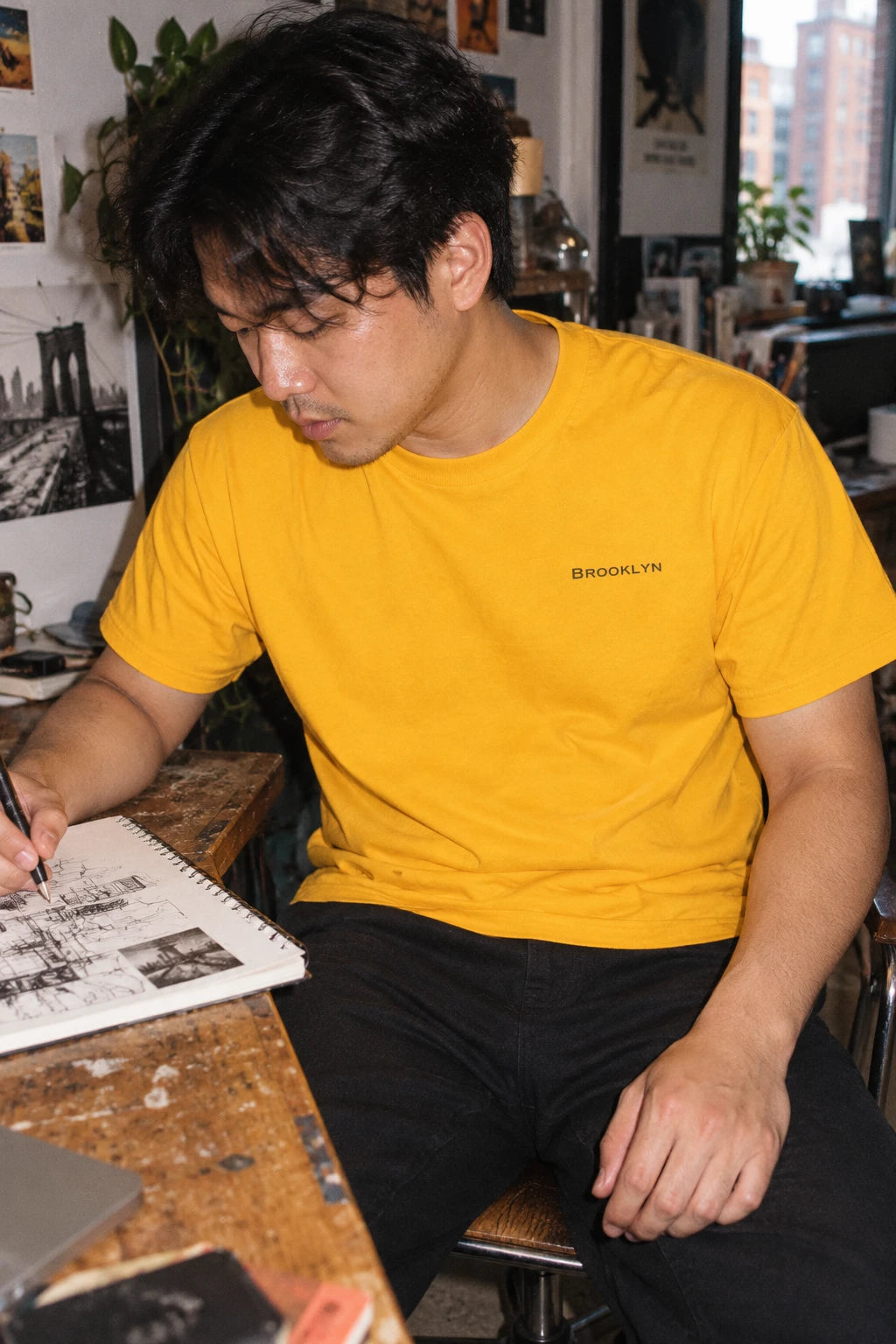 Man wearing a golden yellow Brooklyn T-shirt while sketching at a wooden studio desk.