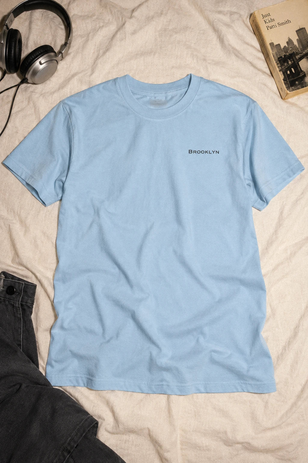 Light blue Brooklyn T-shirt laid flat on ivory bedding beside headphones, dark trousers and a book.
