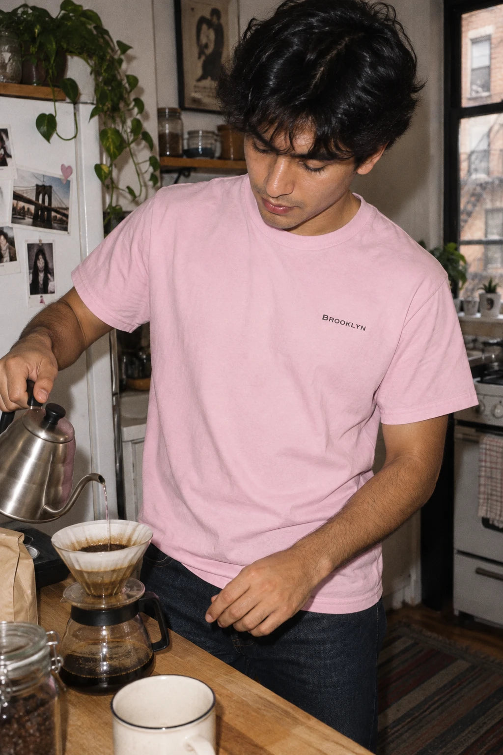 Man wearing a pink Brooklyn T-shirt with small black chest lettering while making pour-over coffee.