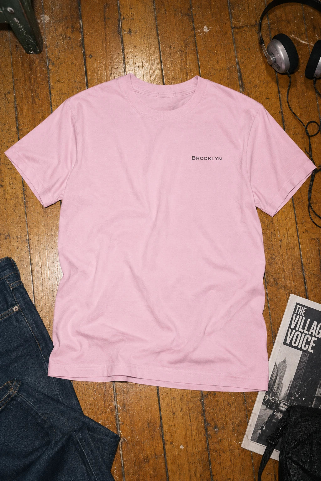Pink Brooklyn crewneck T-shirt laid flat on a wooden floor beside jeans, headphones and a magazine.