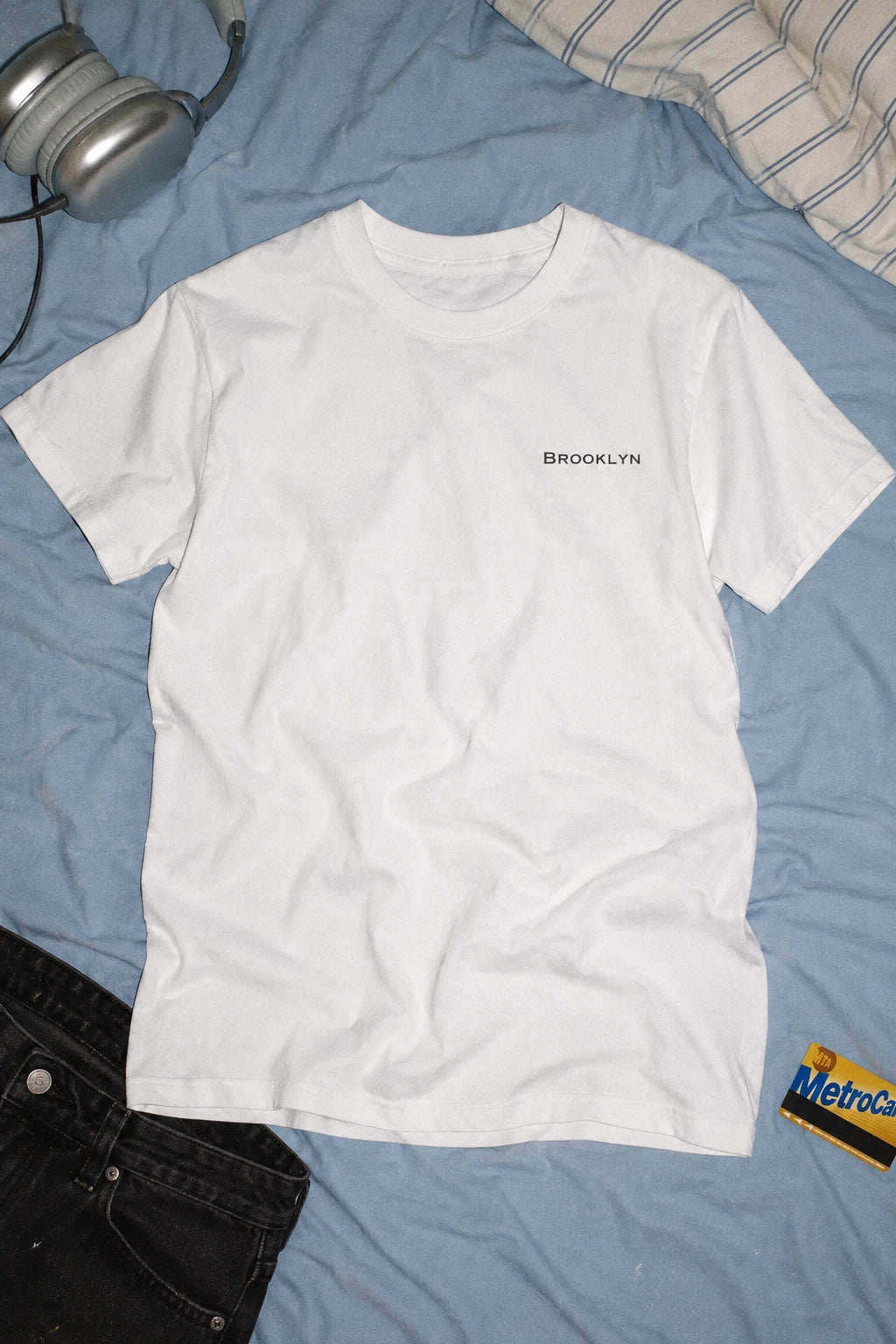 White Brooklyn T-shirt laid flat on blue bedding beside headphones, black jeans and a MetroCard.
