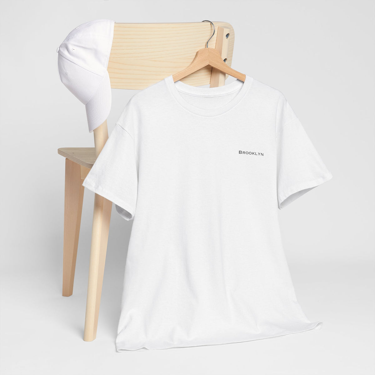 White Brooklyn unisex T-shirt on a wooden hanger in front of a light wood chair.