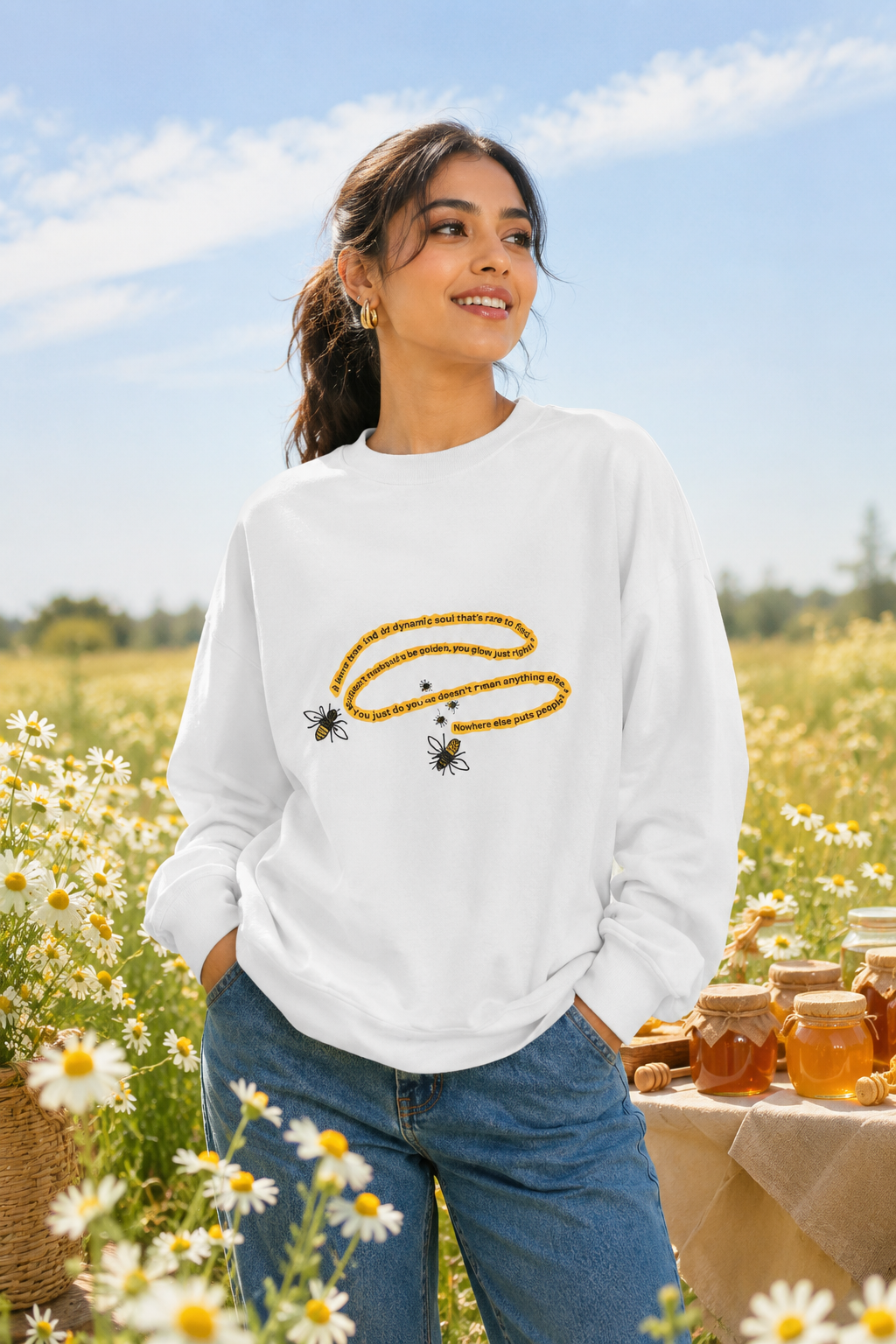 Woman wearing a white bumblebee sweatshirt in a sunny daisy field with honey jars.