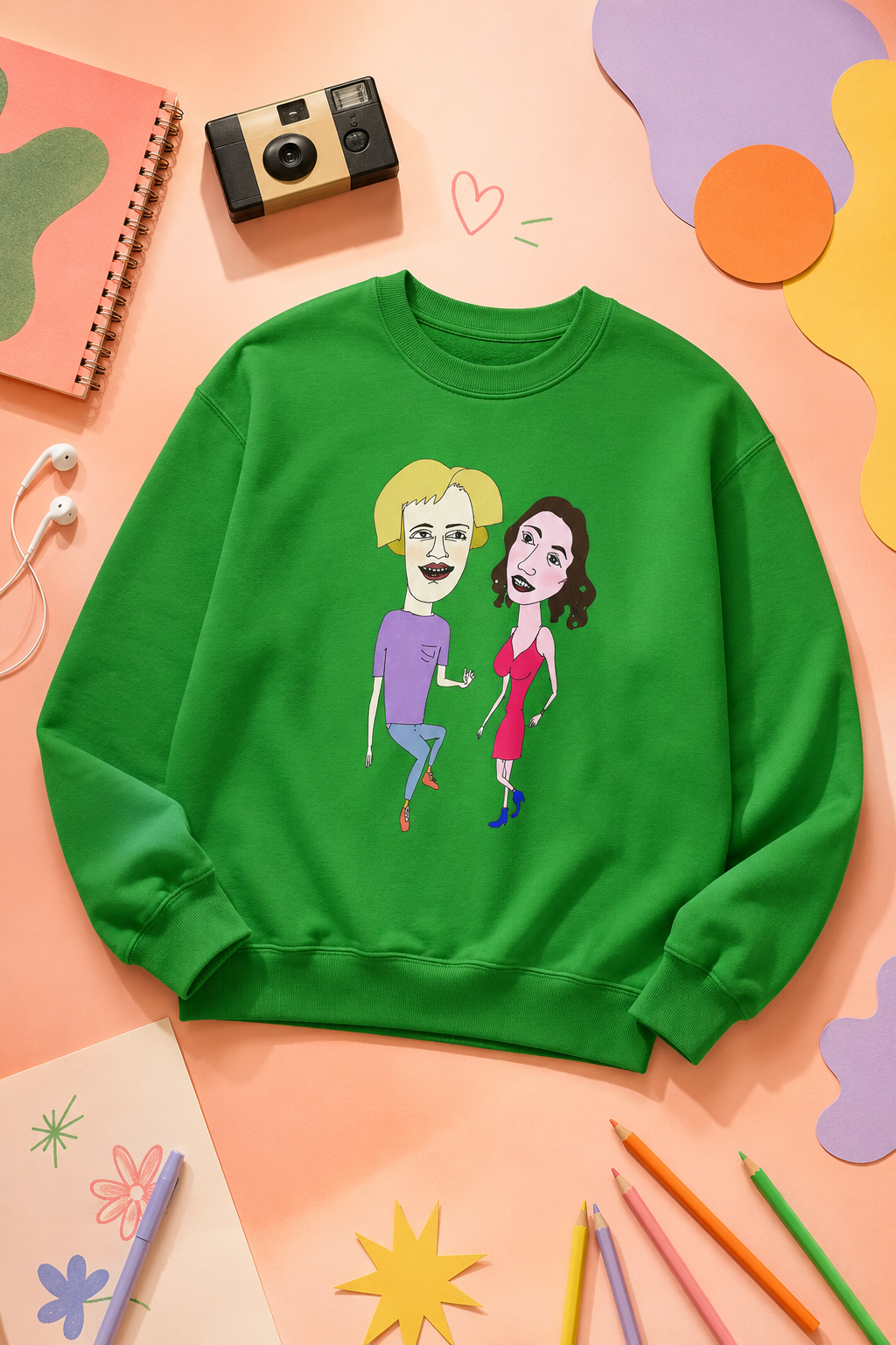 Green cartoon couple graphic sweatshirt flat lay on a pastel peach background with colorful artsy accessories