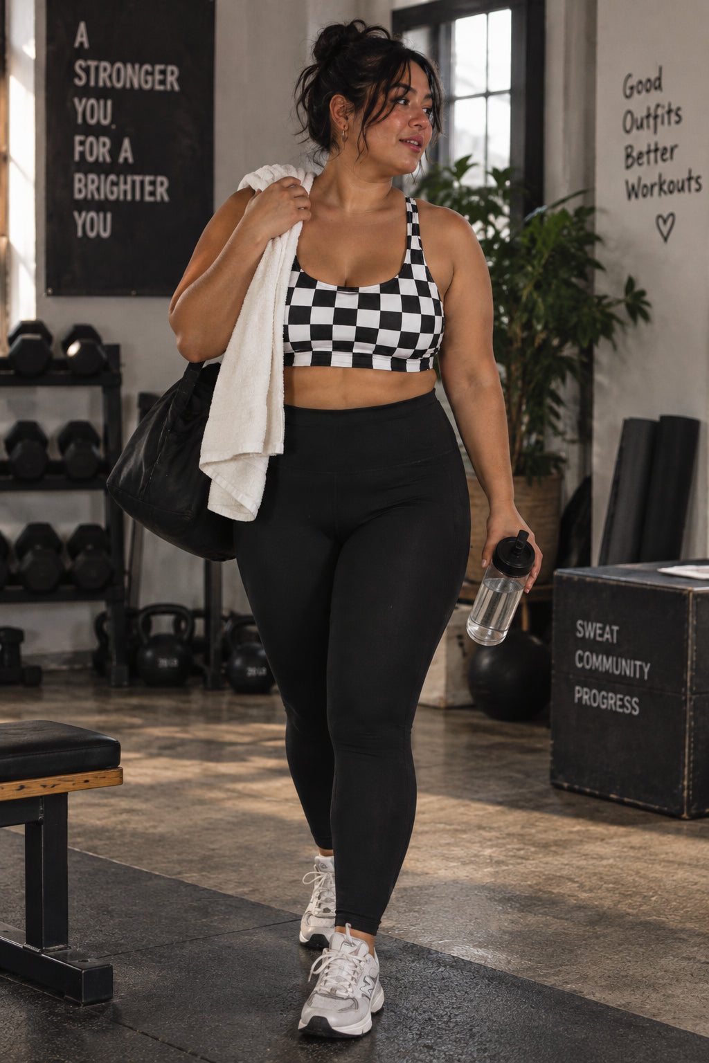 Curvy woman wearing a black and white checkerboard sports bra with high-waisted black leggings, carrying a gym bag, towel and water bottle.