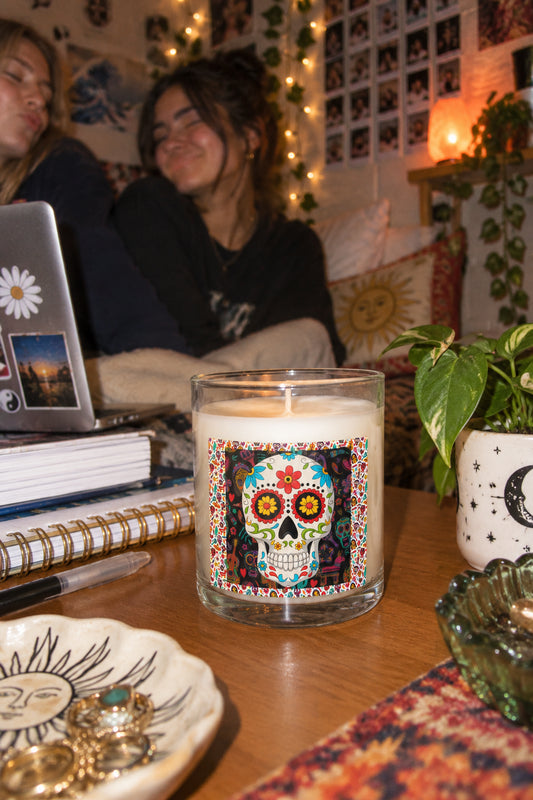 Day of the Dead sugar skull candle on a cozy dorm room desk with books, laptop, plants and warm ambient lighting.