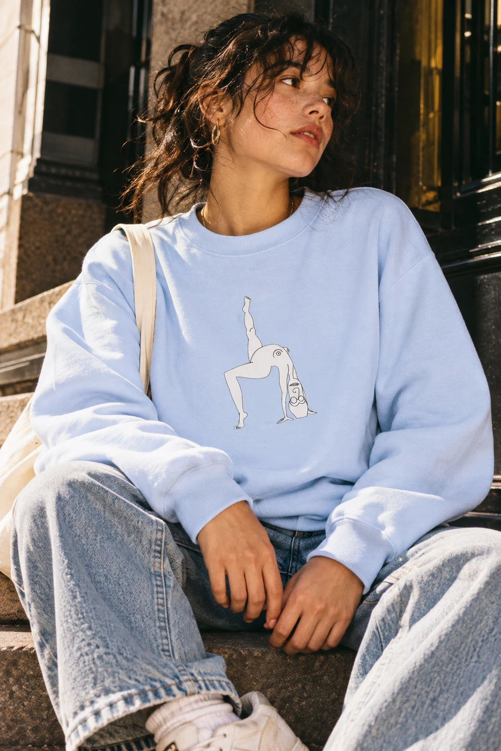 Young woman wearing the Hydrangea blue Elastika Girl graphic sweatshirt while sitting on outdoor city steps.