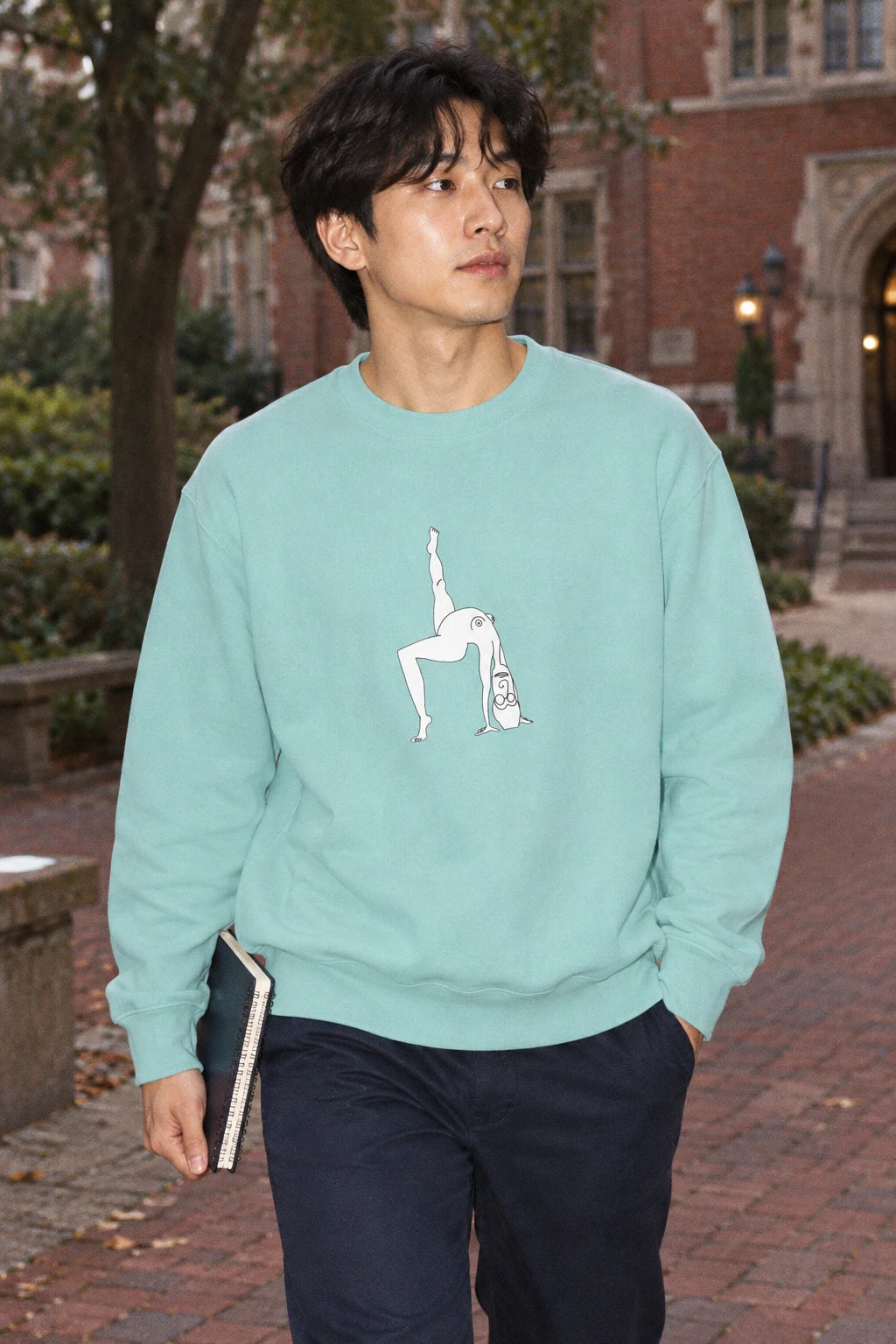 Young man wearing a Chalky Mint Elastika Girl graphic sweatshirt with white line art on a brick-lined walkway.