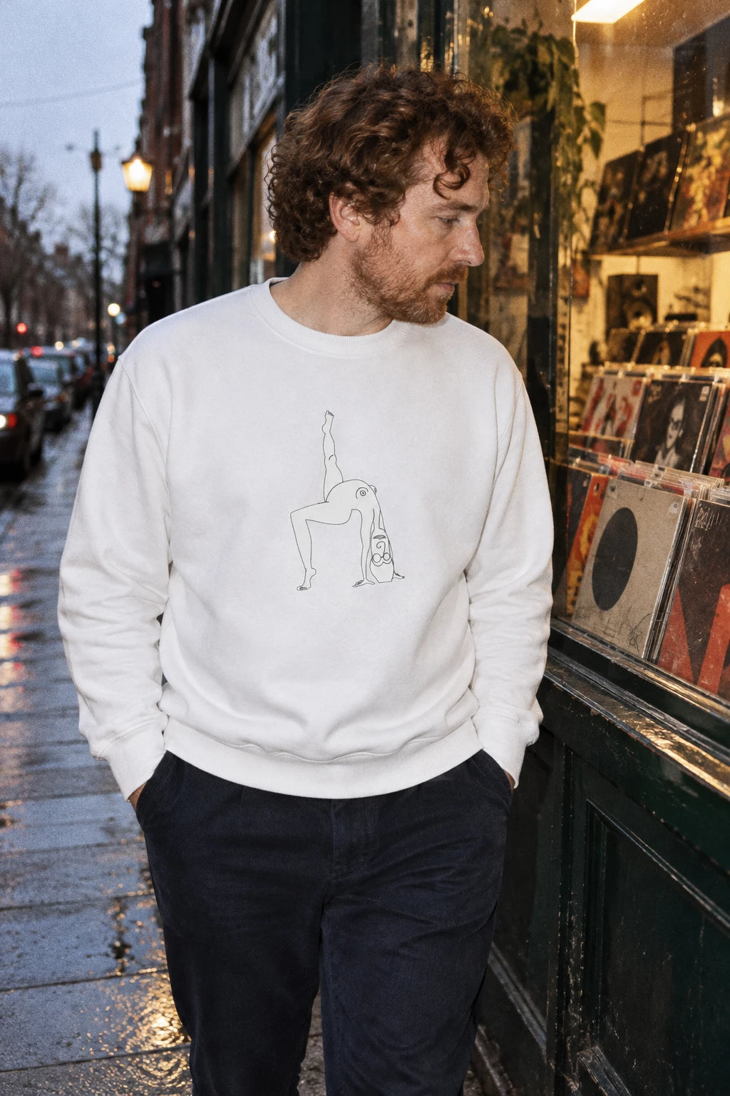 Man wearing a white Elastika Girl graphic crewneck sweatshirt with minimalist line art on a city street.