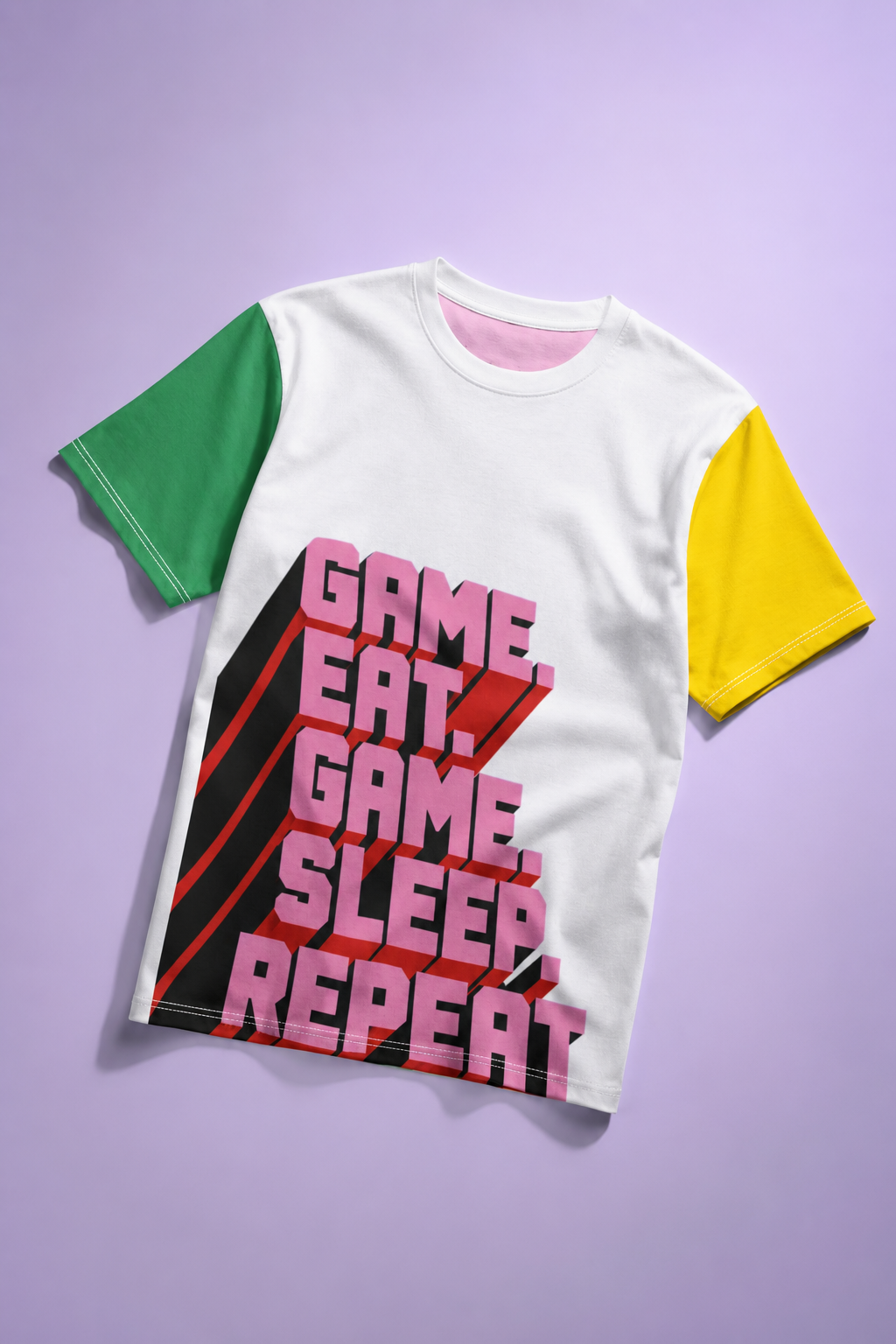 White gamer graphic T-shirt flat lay on lavender background with green and yellow sleeves and bold pink text reading “Game Eat Game Sleep Repeat.”
