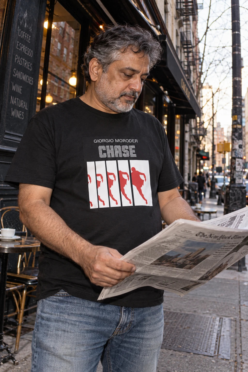 Man wearing a black Giorgio Moroder Chase graphic T-shirt while reading a newspaper outside a city café.