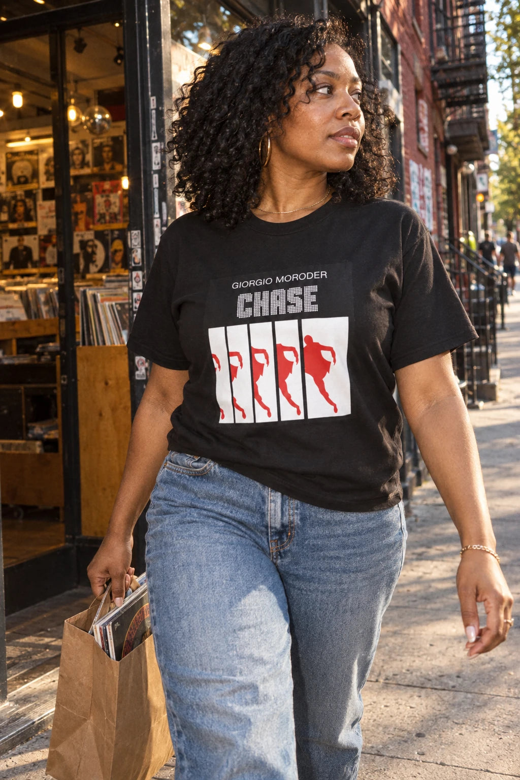 Woman wearing a black Giorgio Moroder Chase T-shirt and jeans while carrying records outside a record store.